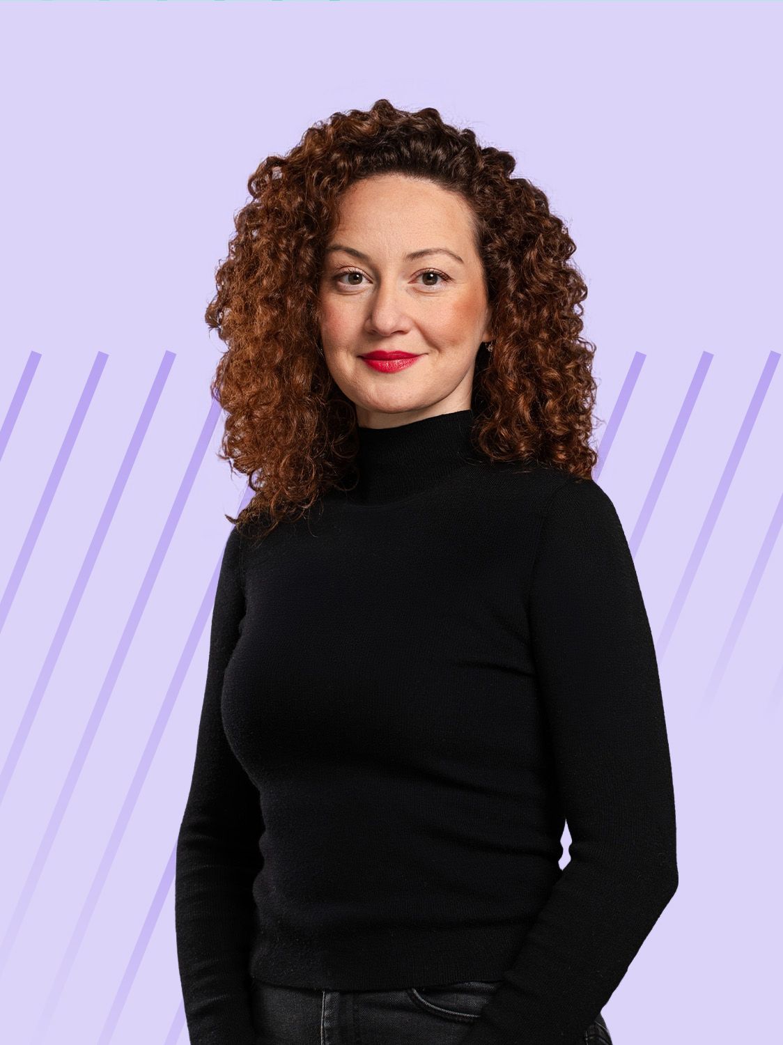 Woman with curly red hair and black turtleneck smiles against a subtle purple background.