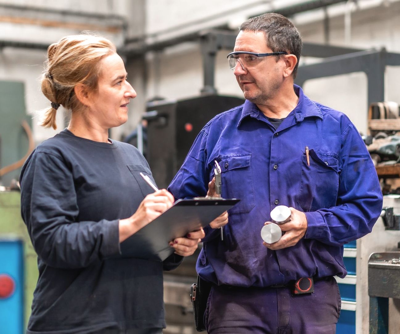Two factory workers discuss production, one holding a clipboard, the other a metal part.