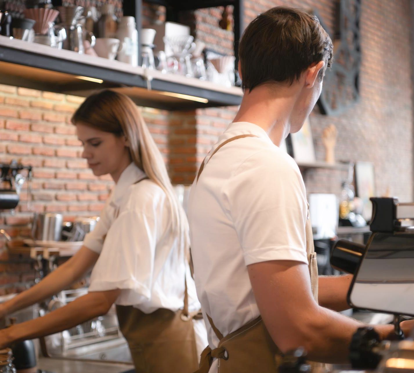 Two baristas prepare drinks behind a modern coffee shop counter with brewing equipment.