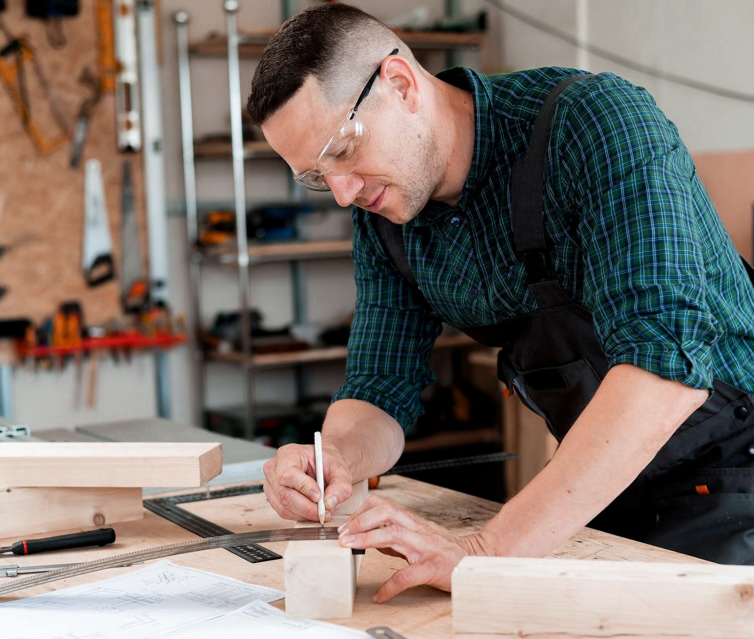 A man in safety glasses precisely marks wood with a pencil in his workshop.