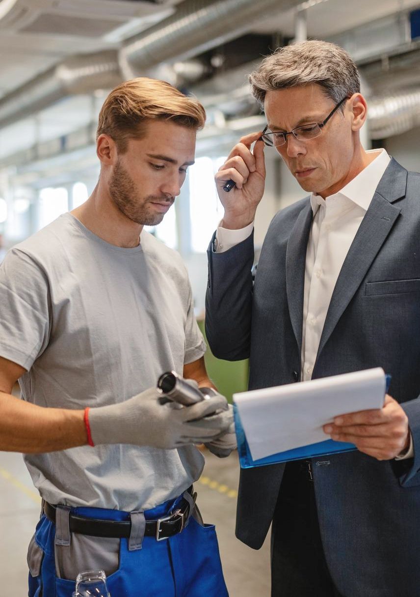 A worker and a manager discuss a small mechanical part in an industrial setting.