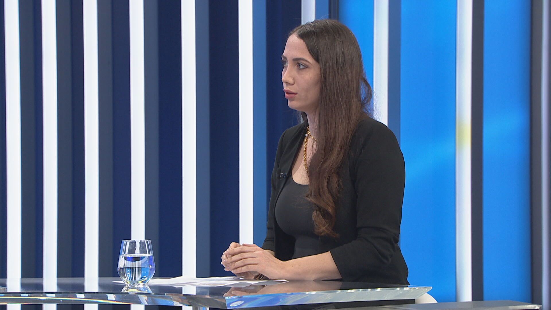 A woman with long dark hair speaks while sitting at a table with a glass of water.
