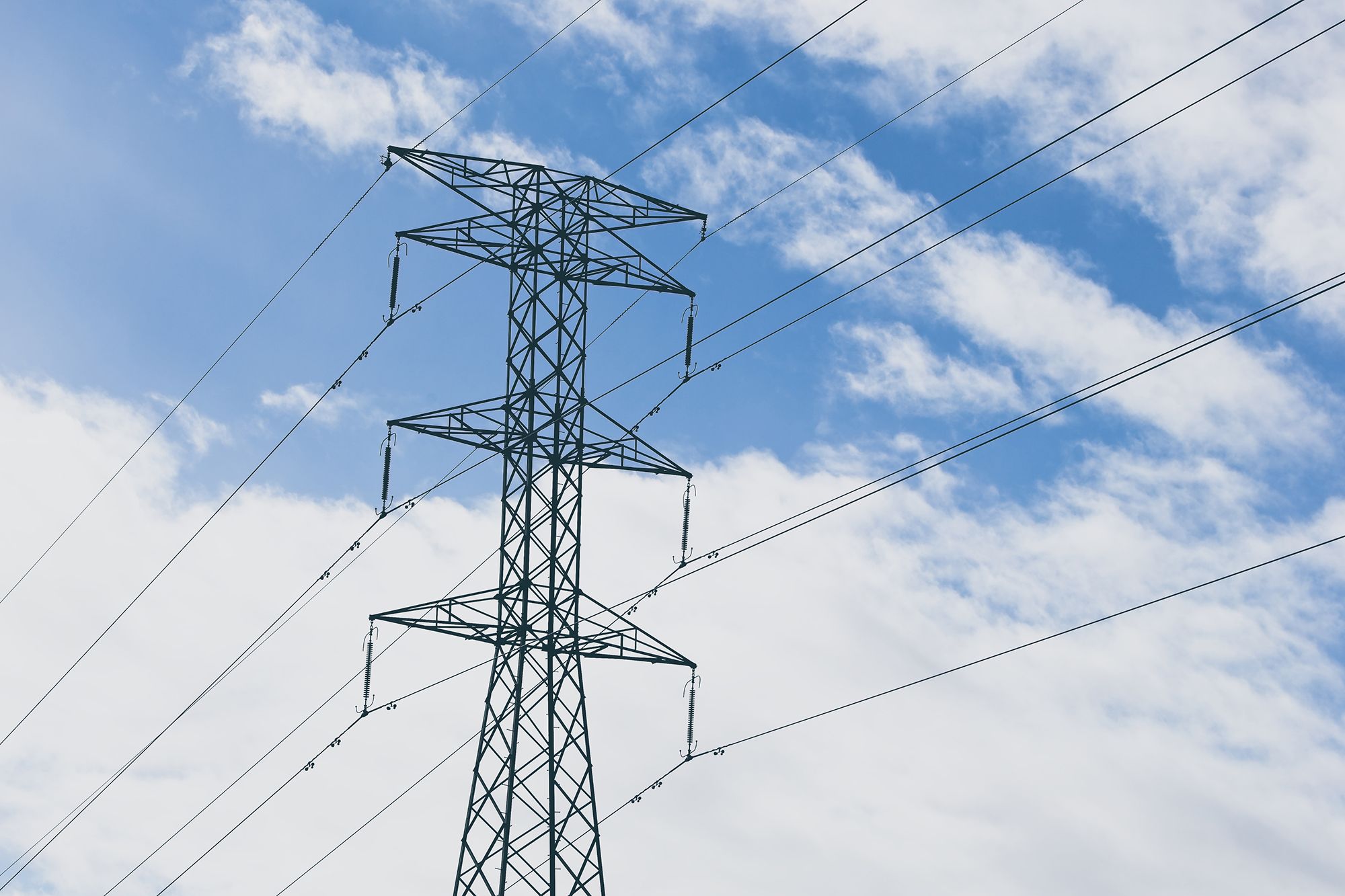 A tall metal electricity pylon stands against a bright blue sky with scattered white clouds.