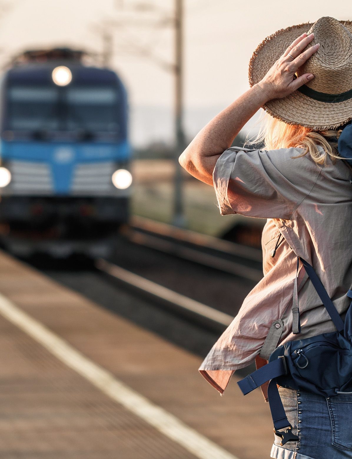 A person in a hat watches a blue train approaching a station platform.