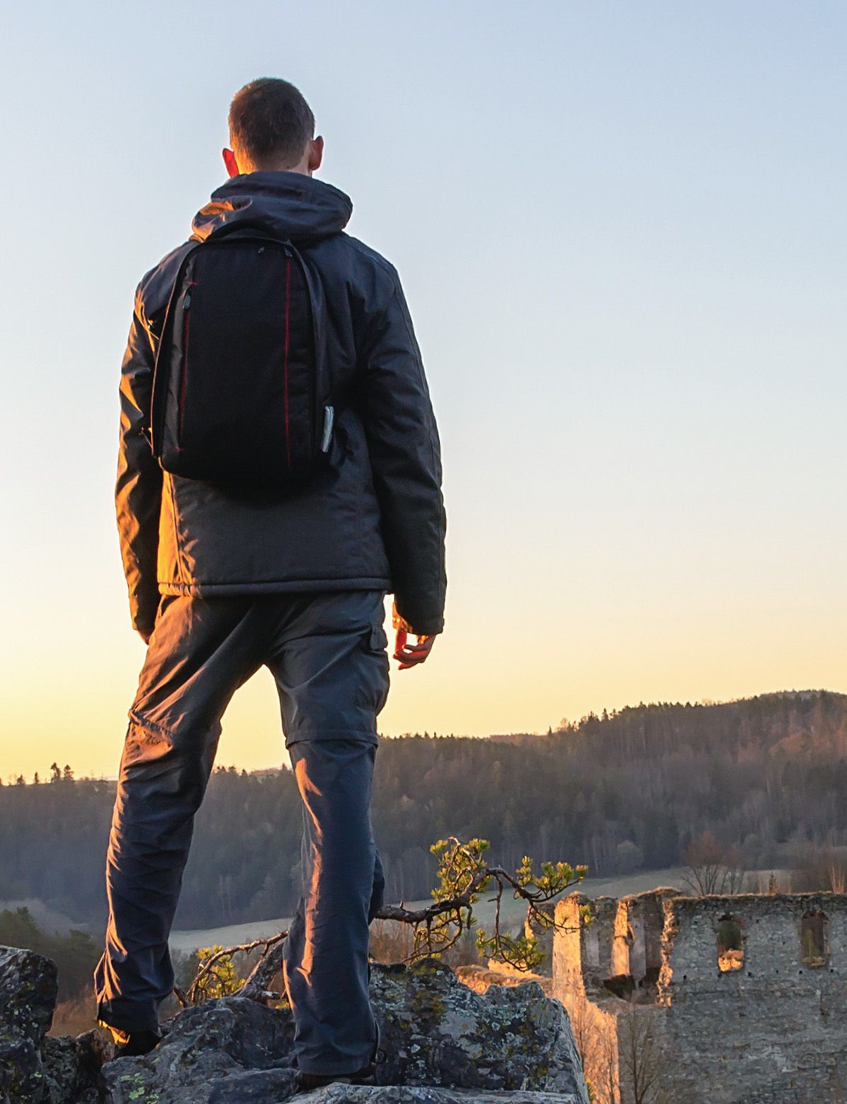 A person with a backpack stands on a cliff, gazing at a mountain sunset.