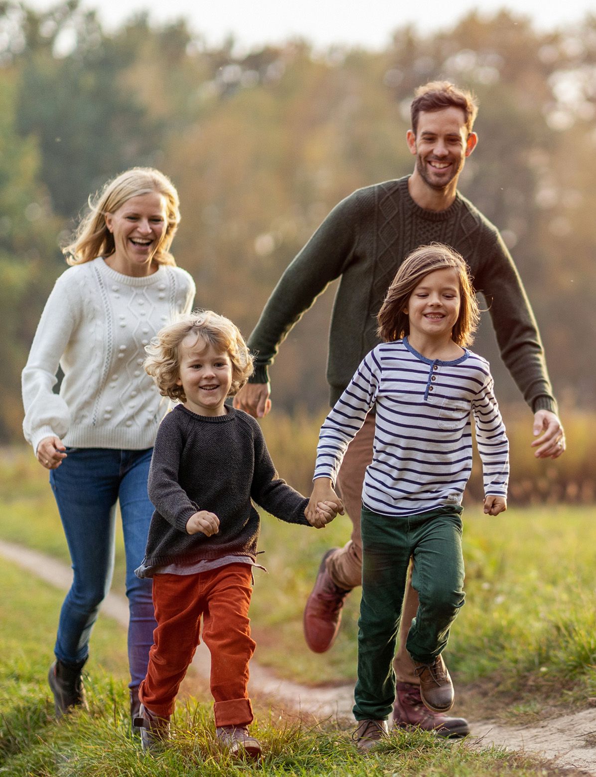 A happy family of four walks hand-in-hand down a sunny outdoor path.