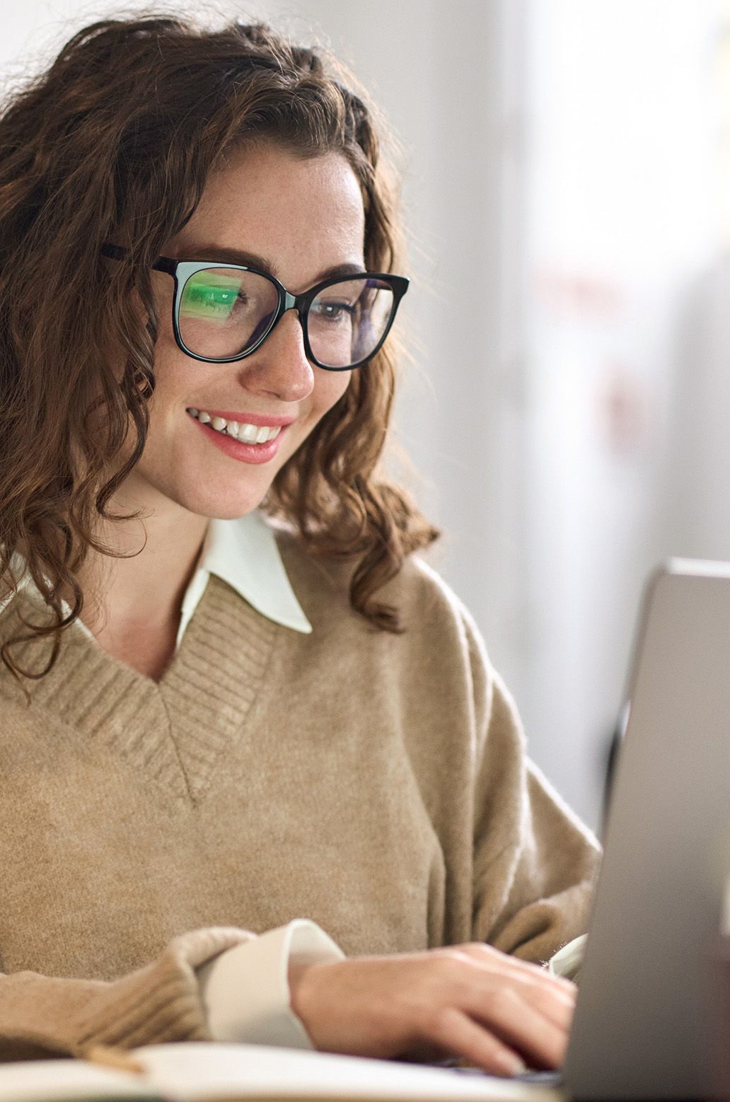 Smiling young woman with curly hair and glasses working on a laptop.