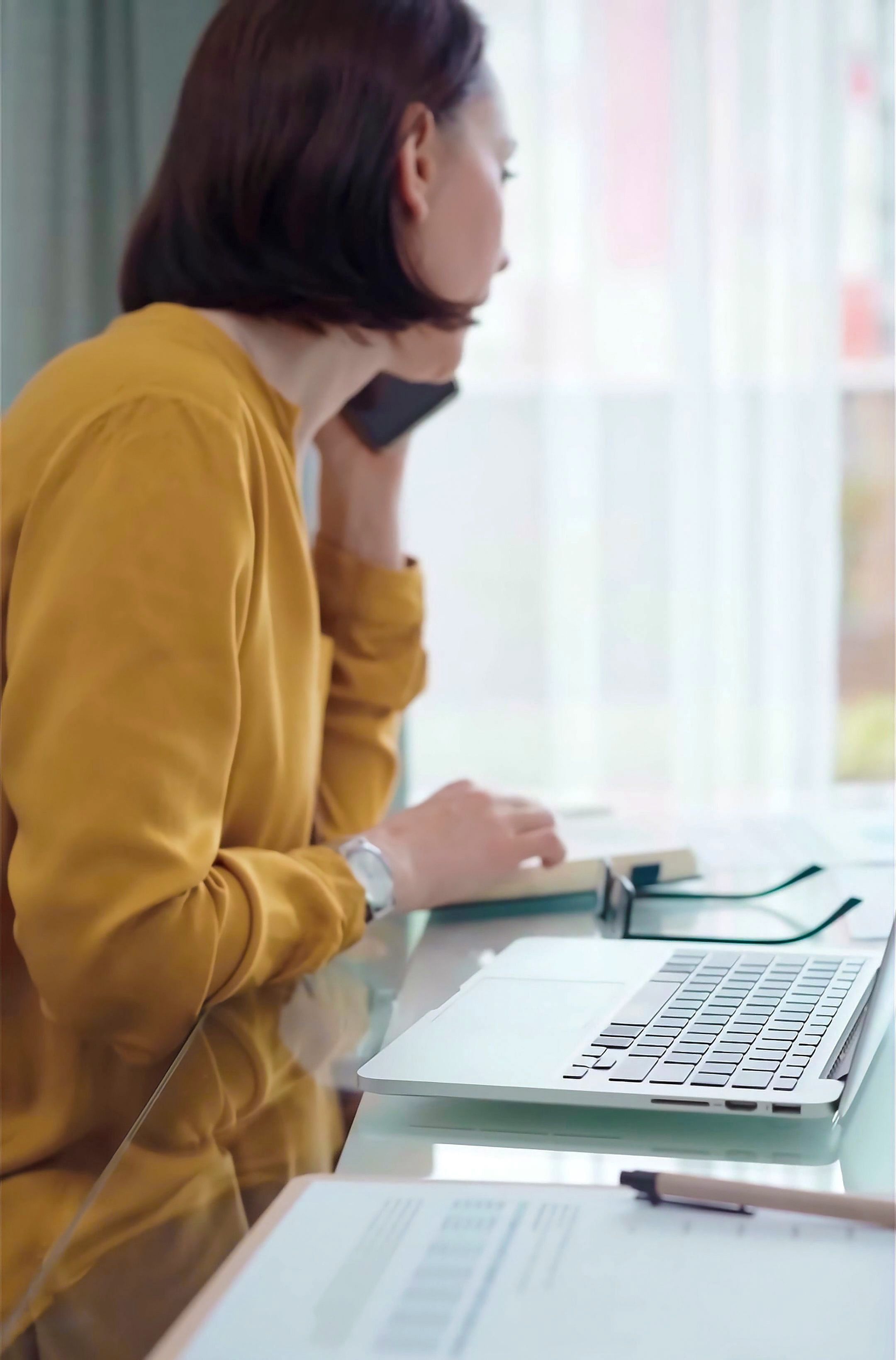 Woman in yellow on phone at a home office desk with laptop.
