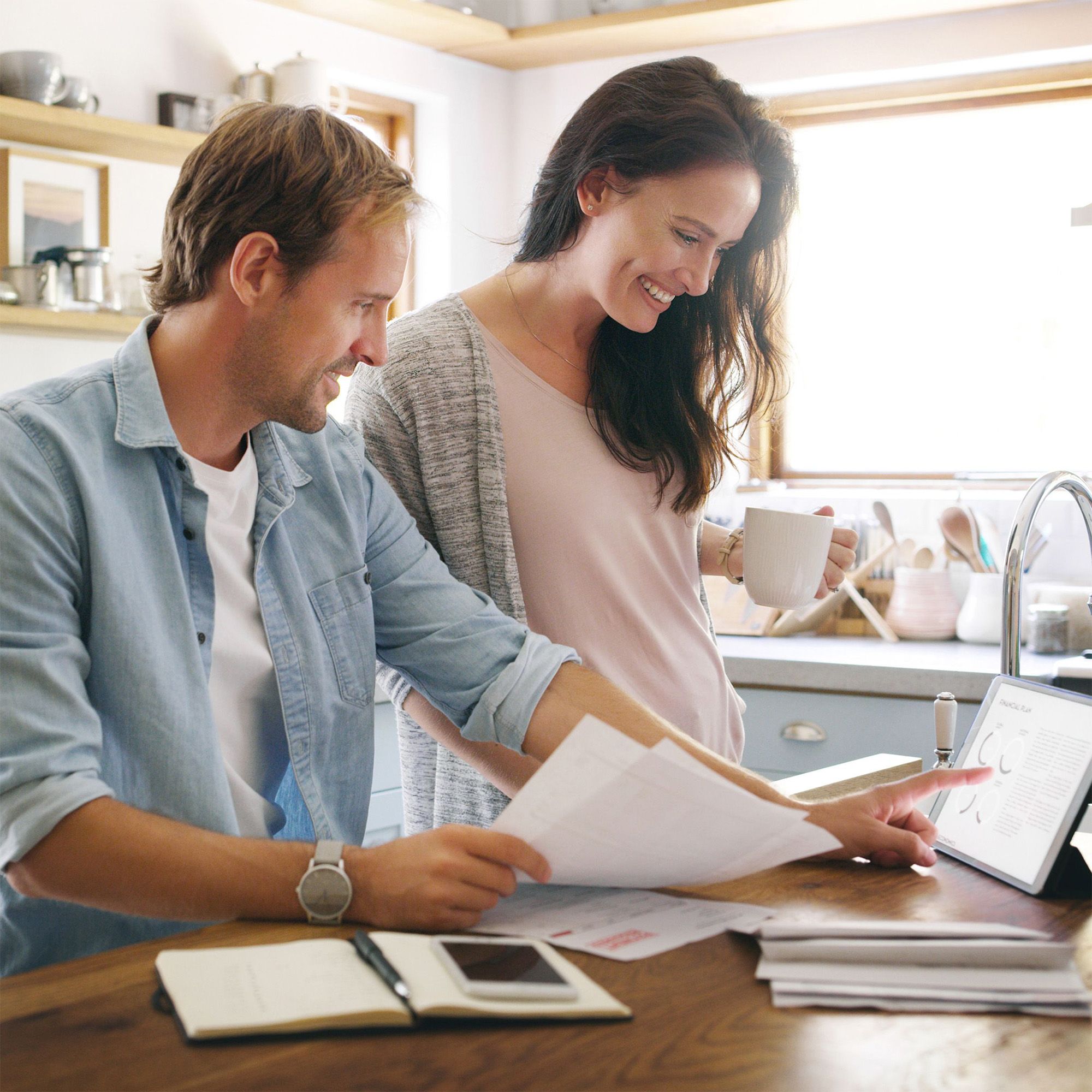A smiling couple reviews documents and a tablet together in a bright kitchen.