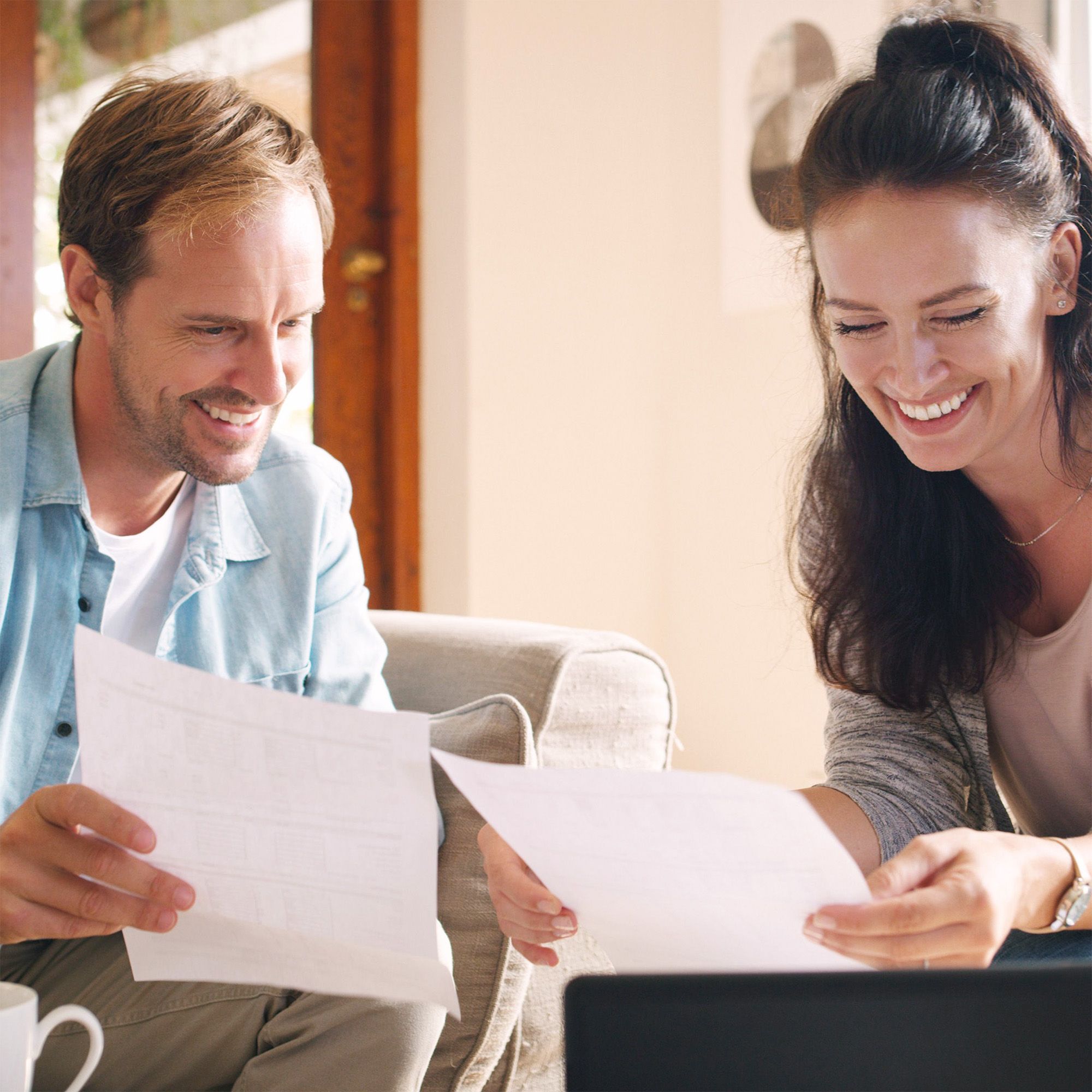 A happy couple smiling while reviewing financial documents and using a laptop at home.