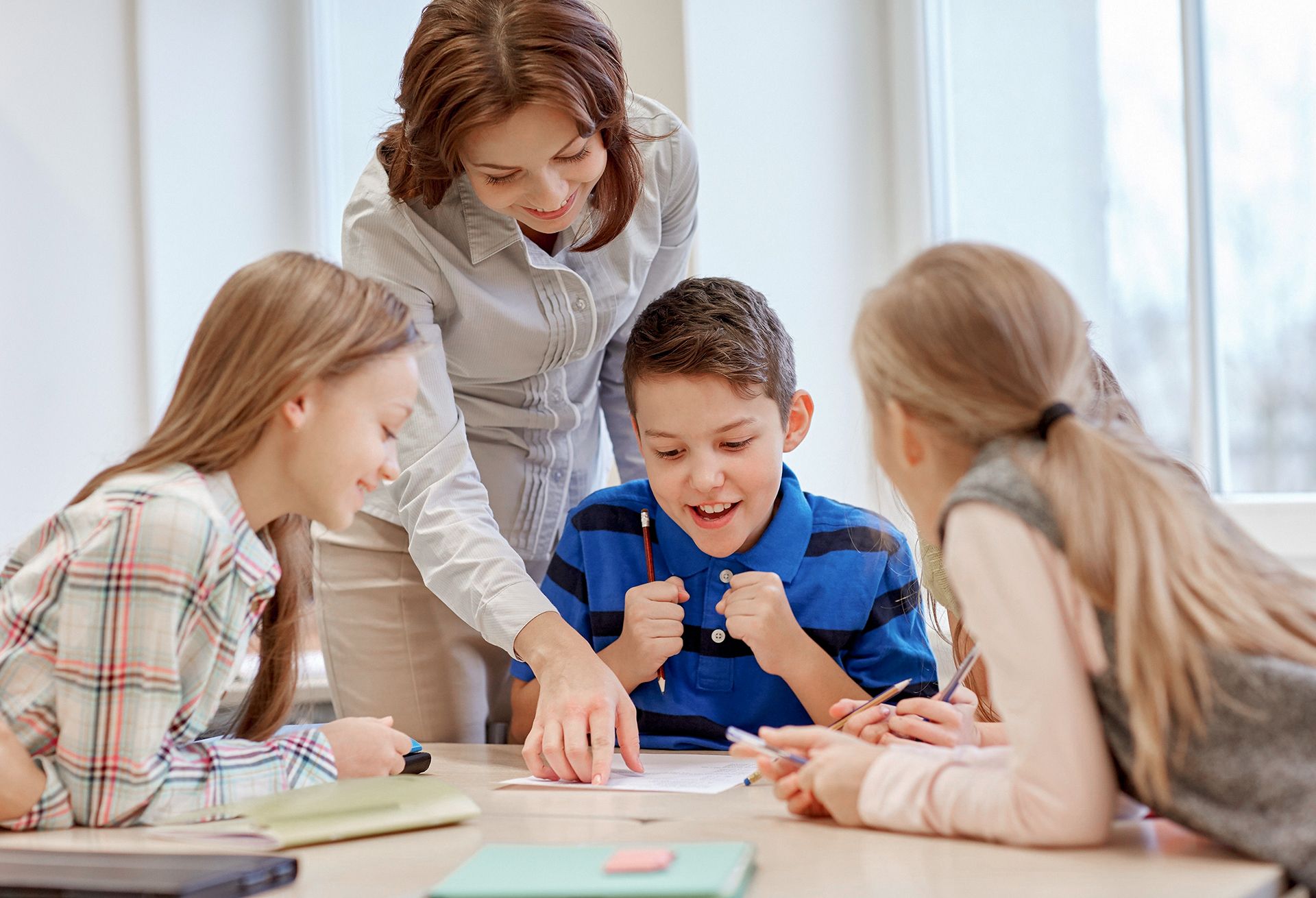 A female teacher helps three smiling elementary school children with a school project.