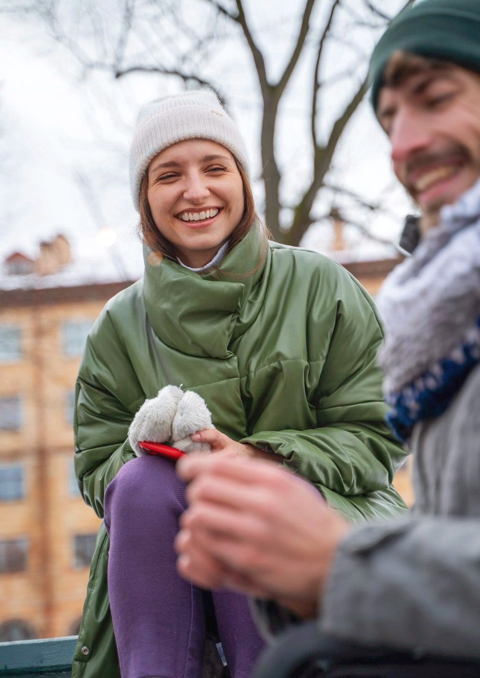Smiling woman in white beanie and green jacket outdoors with blurred companion.