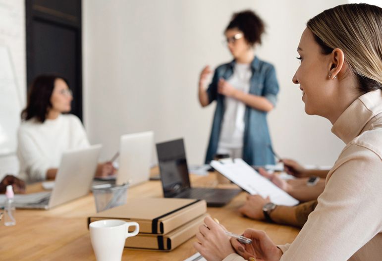 Three women in a meeting, one presenting to two seated colleagues at a wooden table.