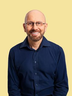 A smiling man with a bald head, beard, and glasses wears a dark blue shirt.