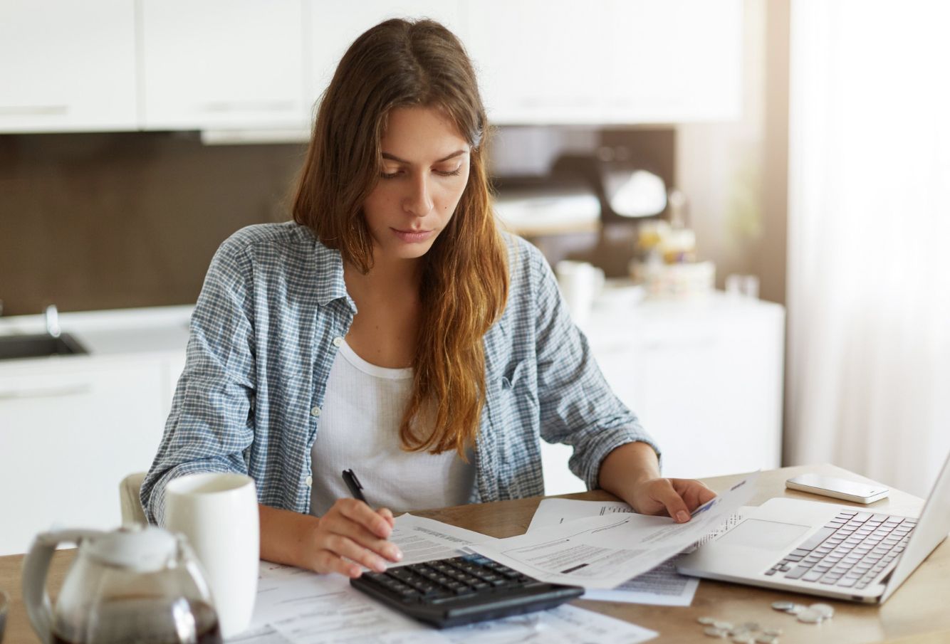 Young woman calculating finances and reviewing documents at a kitchen table.