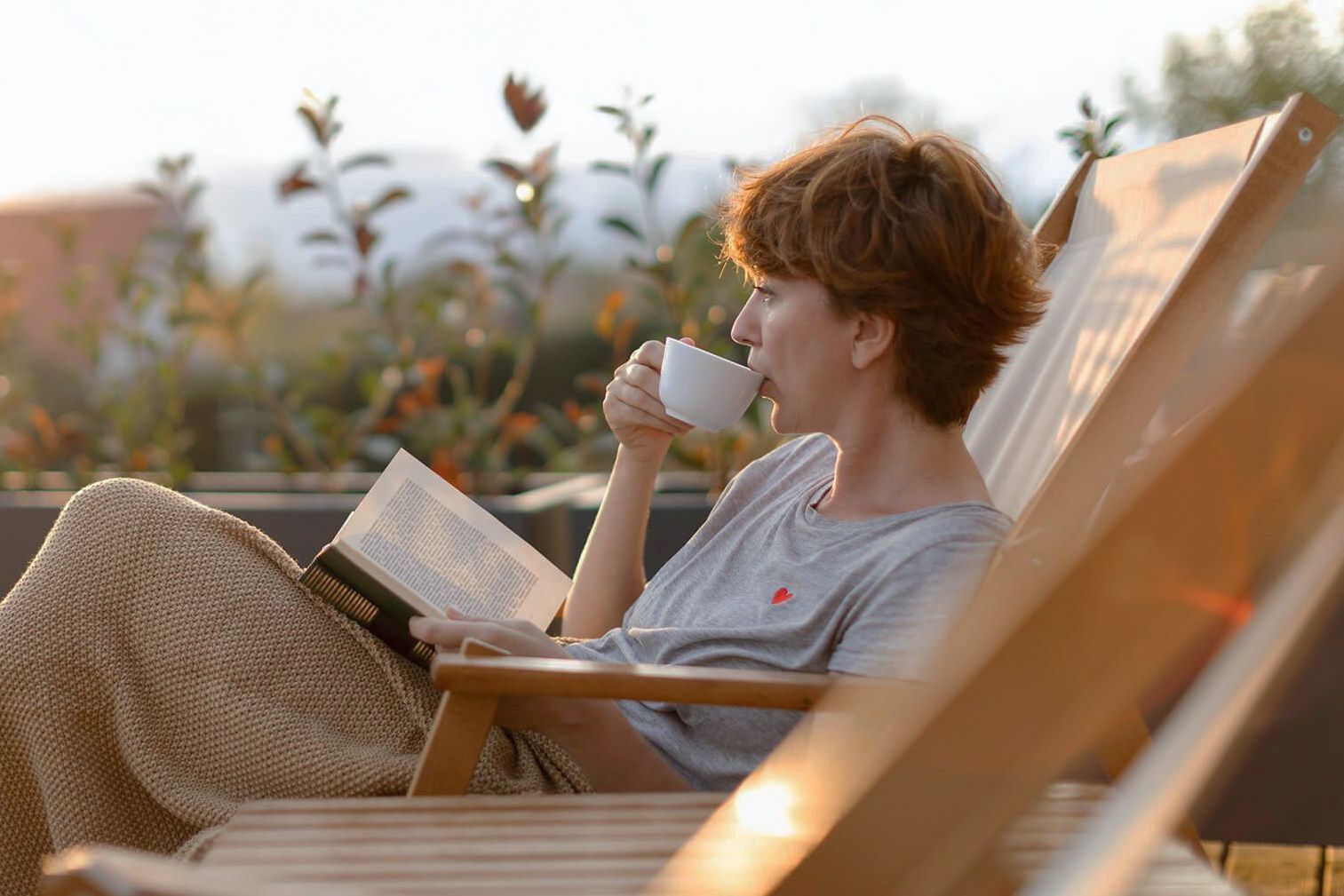 Red-haired woman sips coffee and reads a book on a wooden lounge chair outdoors.