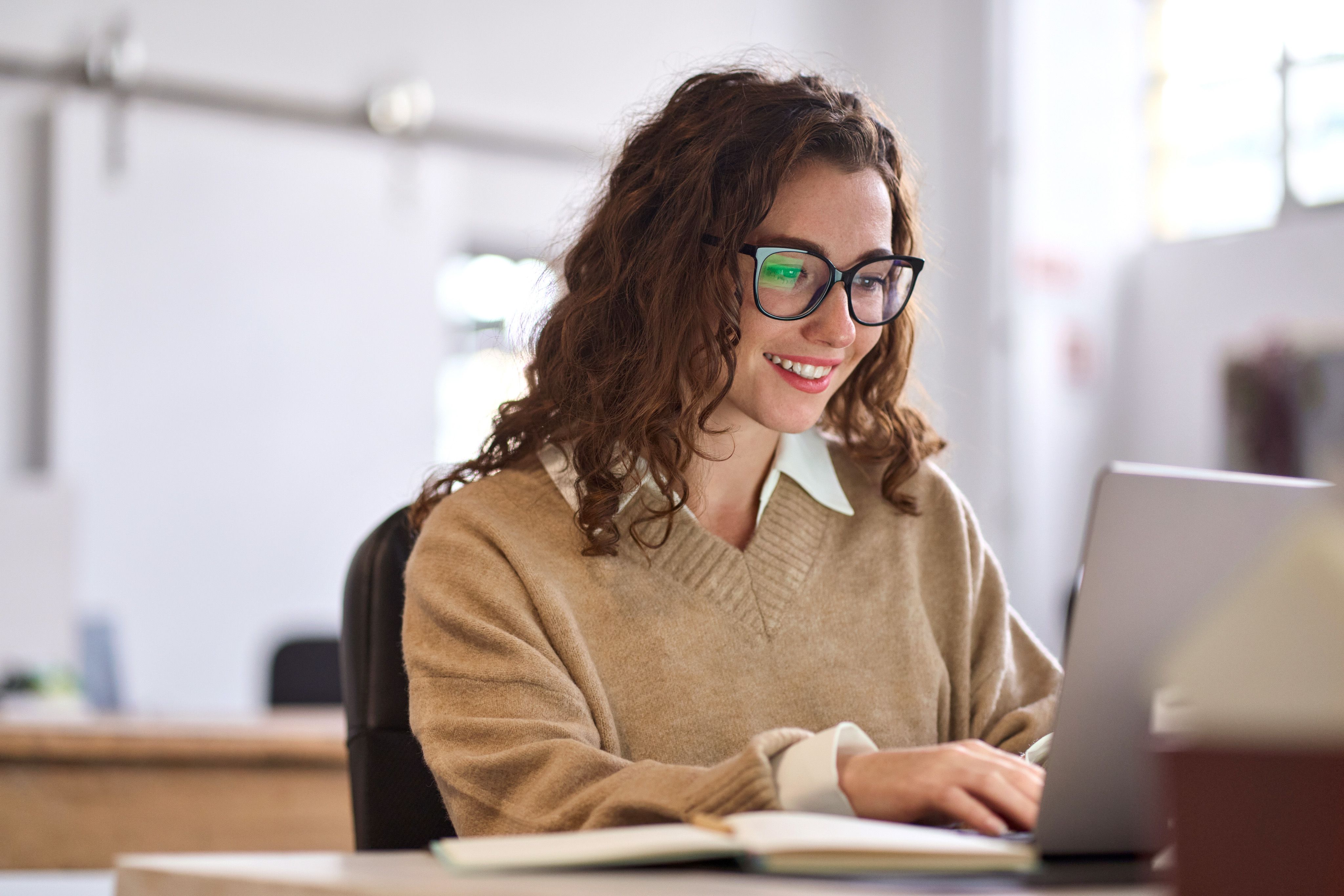 A smiling young woman in glasses works on her laptop.