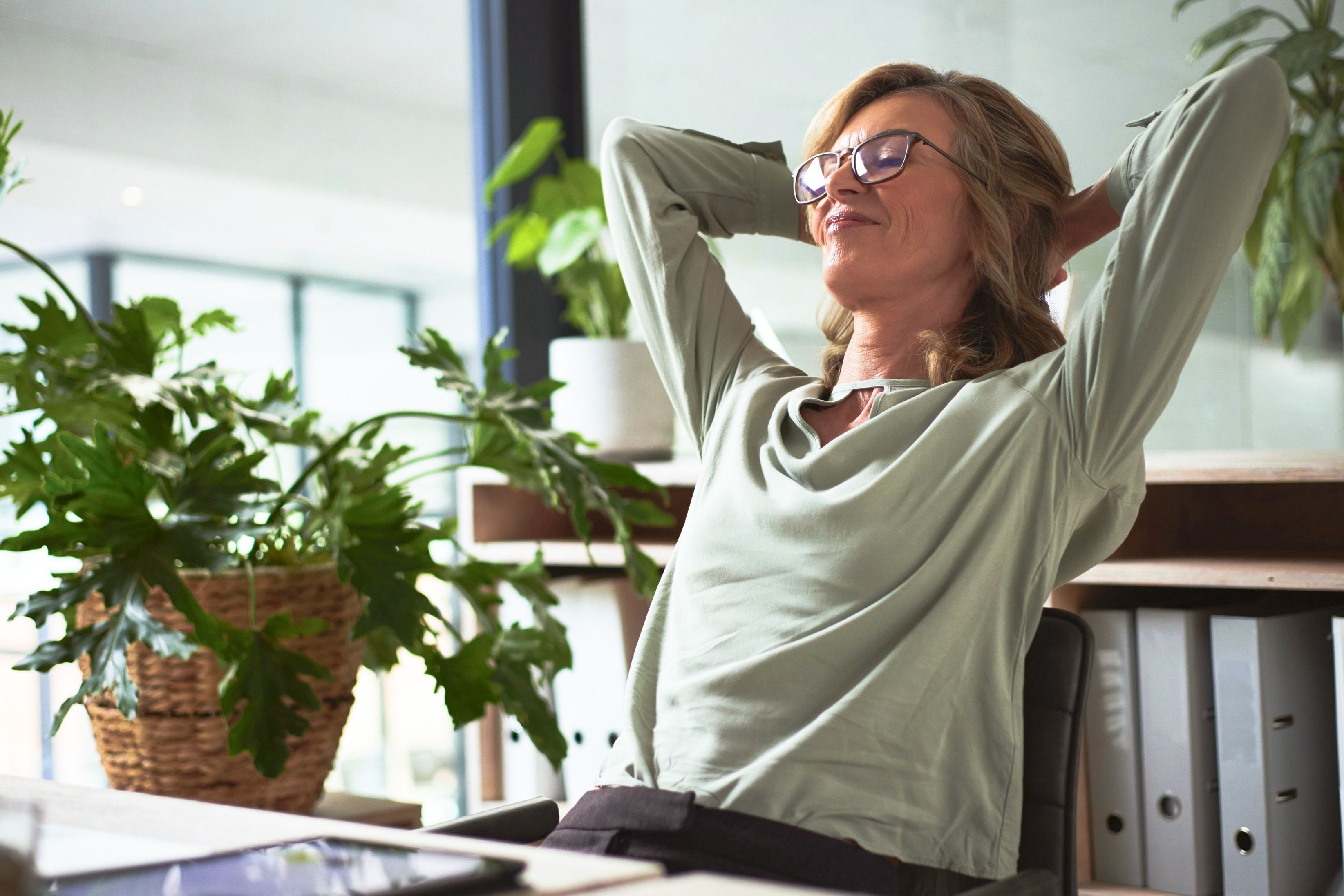 Relaxed woman with glasses leans back in her office chair, hands behind head, eyes closed.