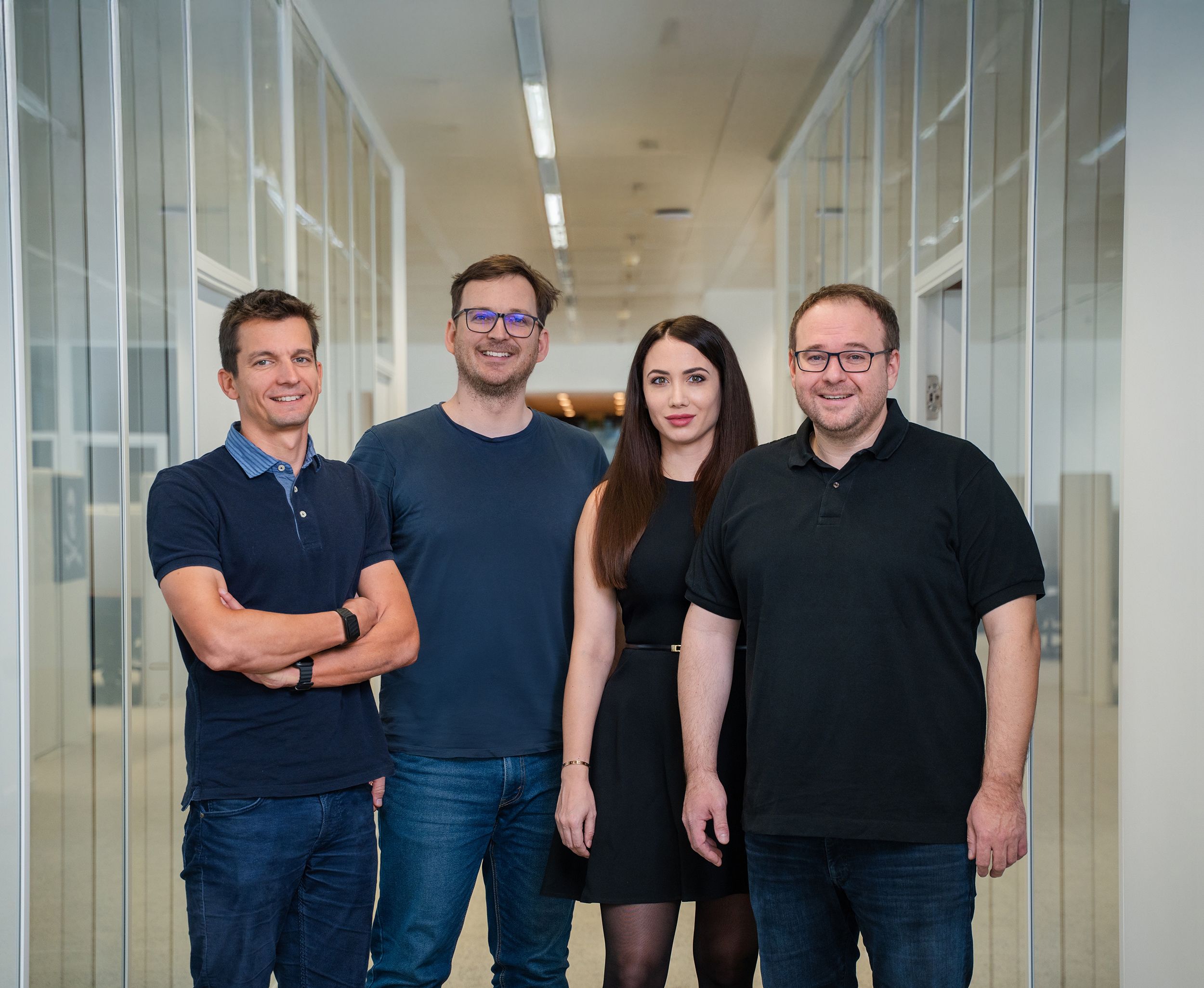 Four smiling colleagues, three men and one woman, stand in a modern office hallway.