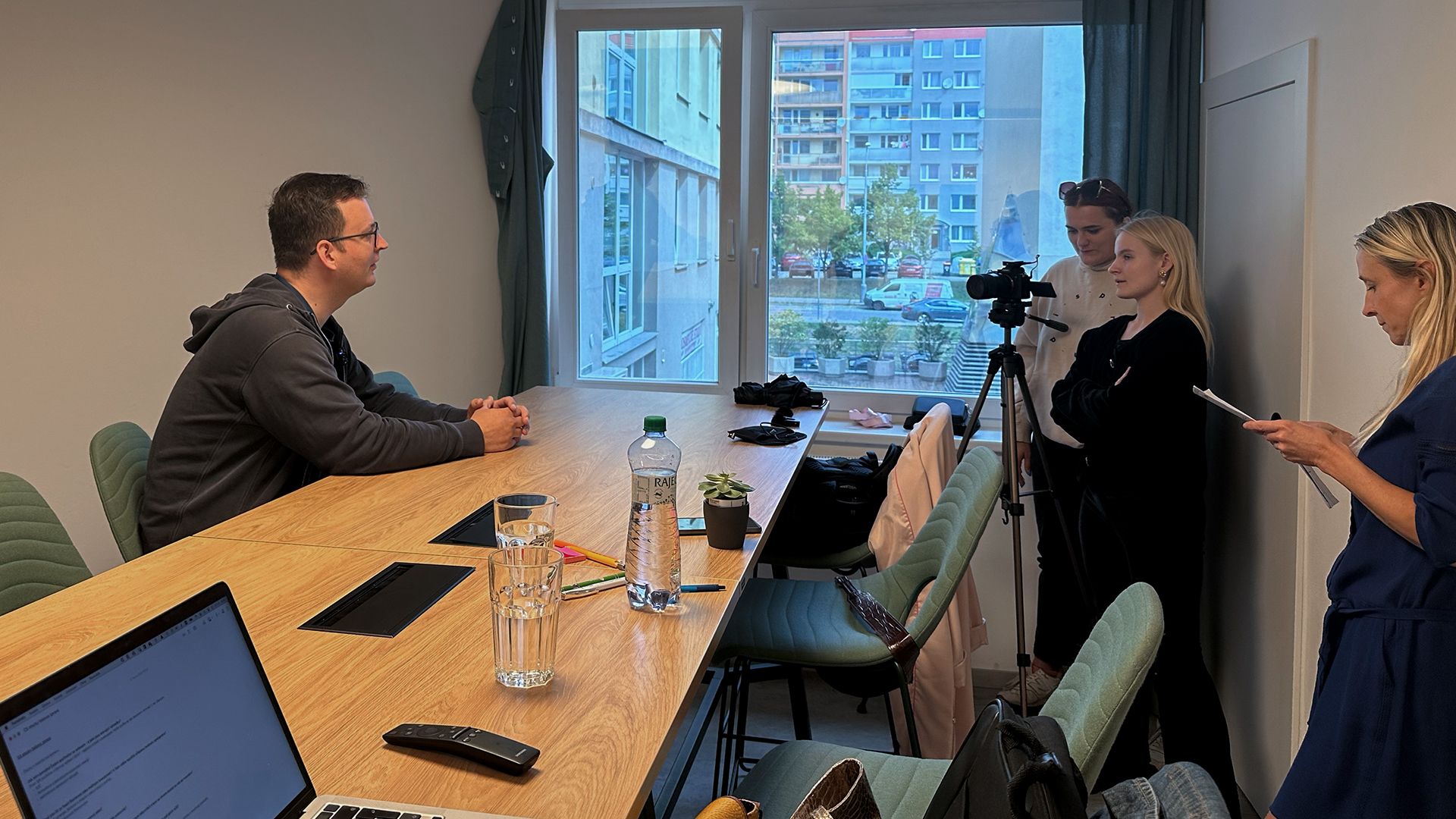 A man is interviewed at a long table, filmed by a crew in an office.