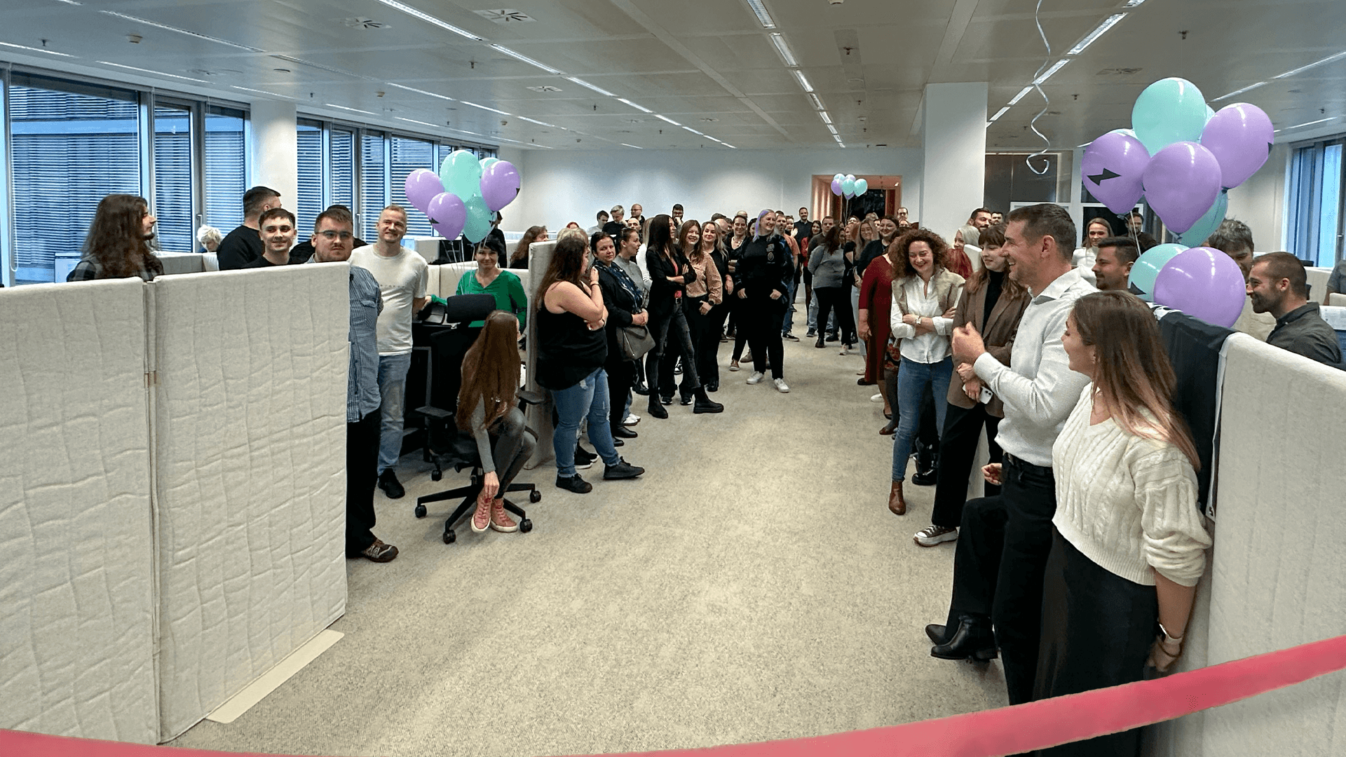 Employees gather in a modern office, celebrating an opening with balloons and a pink ribbon.
