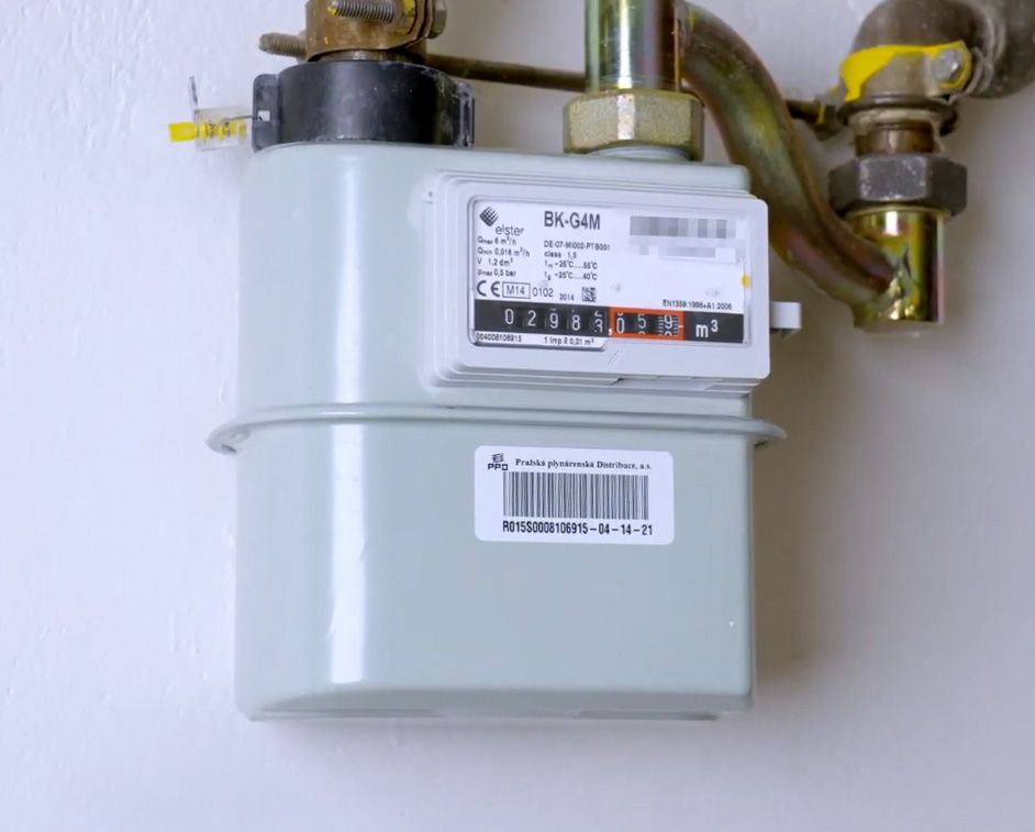 A light grey gas meter with pipes connected, mounted on a white wall.