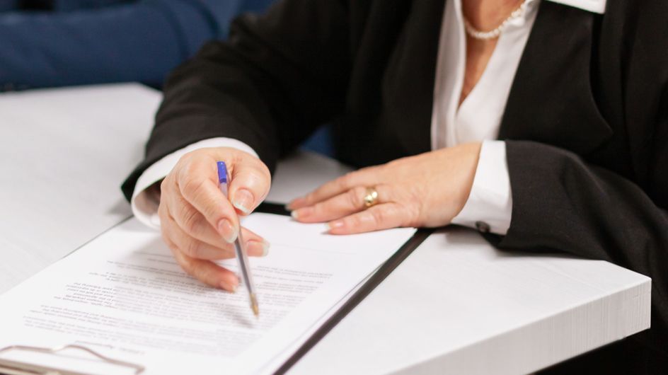Professional signing a document with a pen, wearing a suit and pearl necklace.
