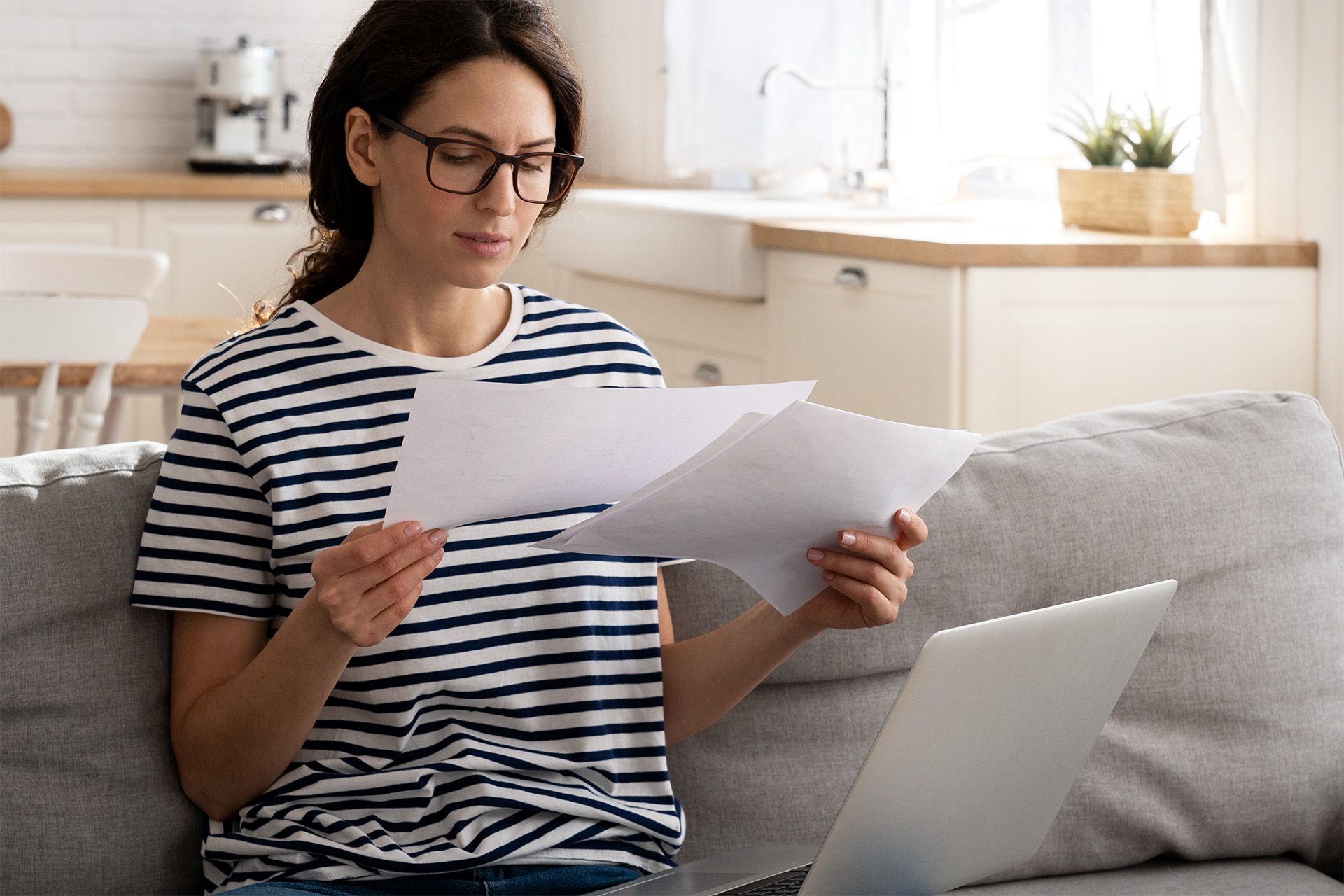 Woman in glasses reviewing documents on a couch with a laptop at home.