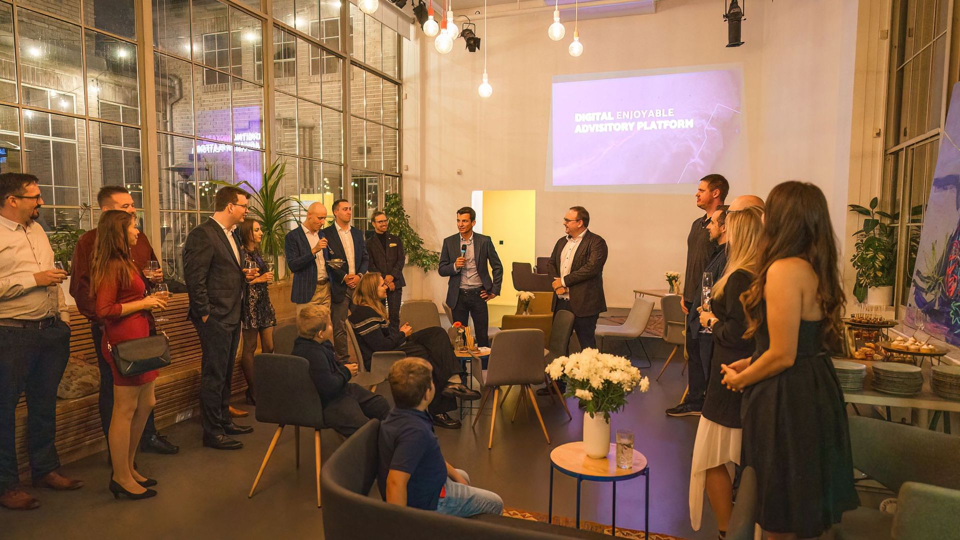 A group of professionals network at an indoor event with a projector presentation.