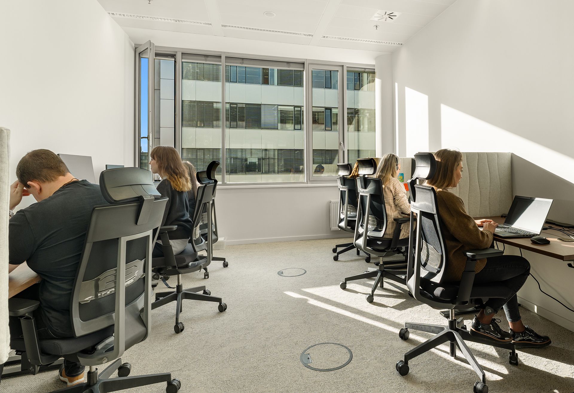 People work at their individual desks in a bright, modern office with large windows.