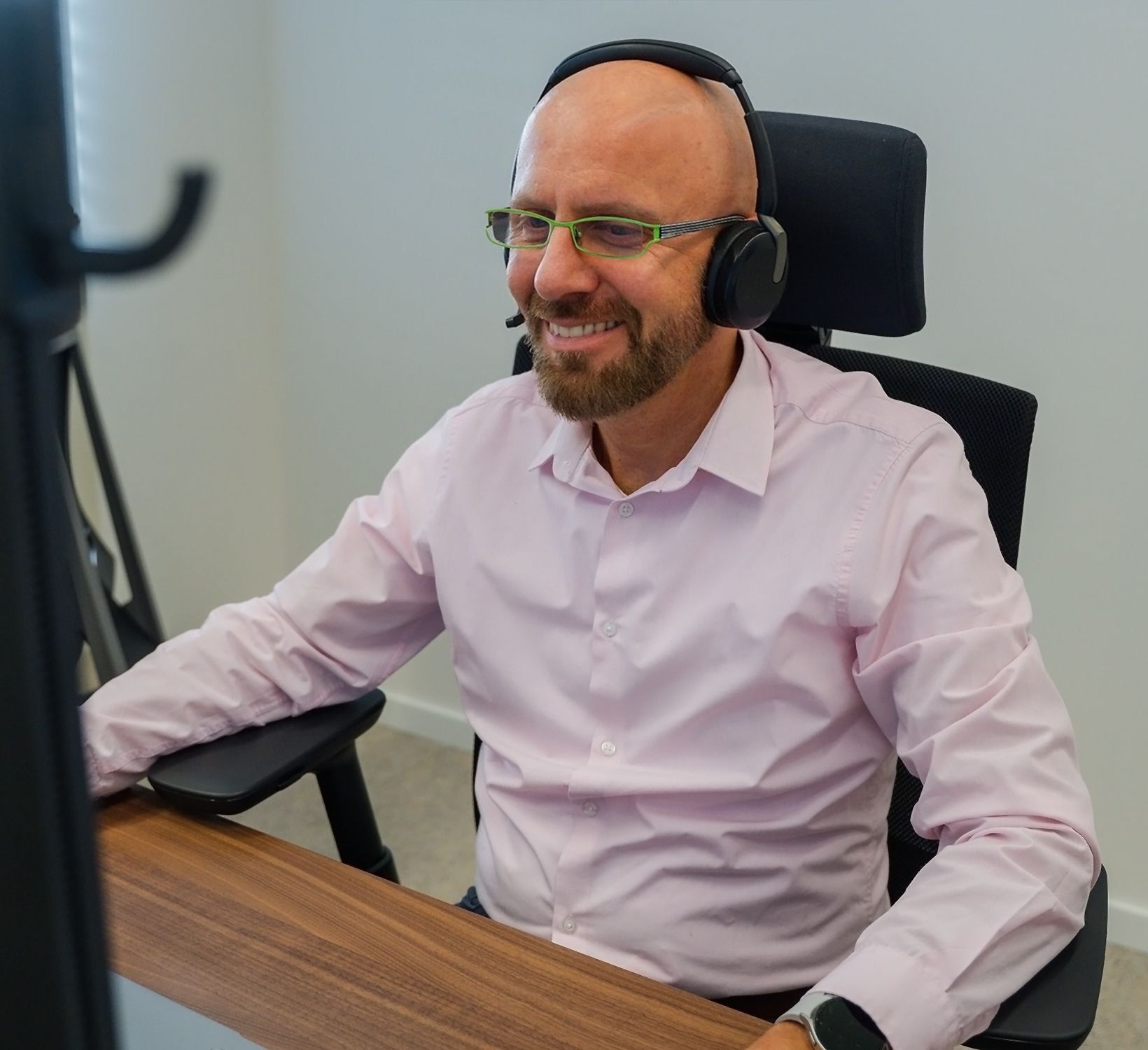 Smiling man with headphones, glasses, and beard working at a wooden desk.
