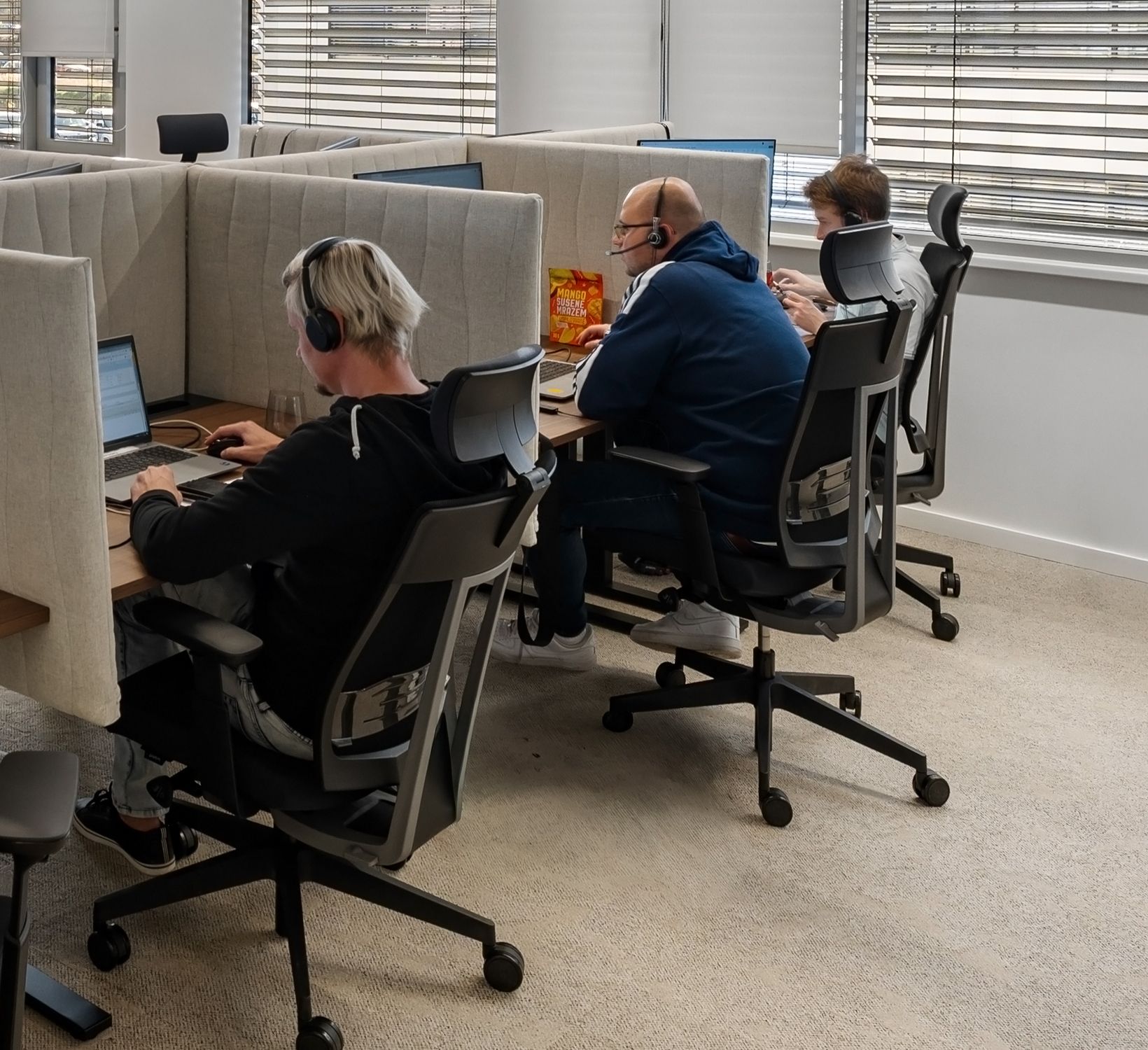 Three people with headphones work diligently on computers in a modern office cubicle setting.